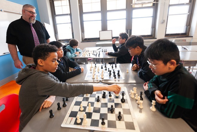 Students seated at tables face each other and play chess while a teacher — a White man in a short-sleeved collared shirt (black) and purple tie, with glasses and a grey beard and grey hair stands, smiles and watches them, part of an after-school time school enrichment program of the School District of Philadelphia.