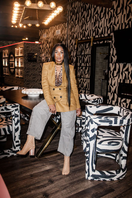 Dominique Calhoun, a Black woman with straight black hair, a gold double-breasted jacket, animal print blouse, modern khakis and beige boots, stands in her design studio among modern armchairs and wallpaper, both with black-and-white print.