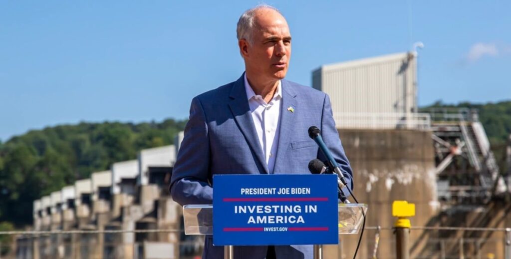 Senator Bob Casey, an older white man with white hair around a bald head wearing a blue suit and white shirt stands outdoors in front of a factory building at a podium with a sign attached to the front that reads, President Joe Biden Investing in America