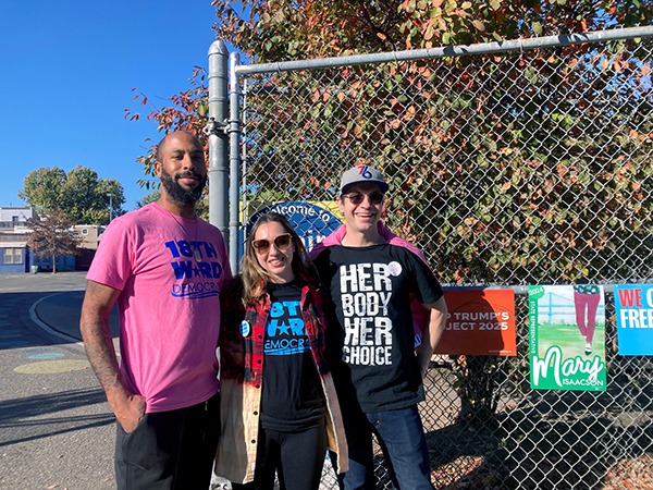 Three people — an African American man with a beard and tattoos on his right arm, wearing a pink t-shirt, a smaller white woman with dirty blond shoulder-length hair wearing a "VOTE HARRIS" t-shirt, jacket and large sunglasses, and another White person in a 76ers cap, sunglasses, and a t-shirt that reads "HER BODY HER CHOICE" and jeans stand outside chain link fence near polling place at Adaire Elementary in Philadelphia. 