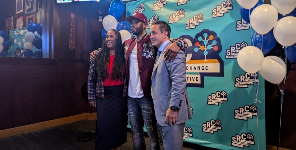 A black woman with long hair a red shit and black skirt stands to the left of a tall black man in a maroon jacket, white t-shirt, and fashionably distressed jeans. On the right is a white man in a light gray suit with salt and pepper hair. Behind them is an aqua blue banner with the logo of the nonprofit SRC printed repeatedly