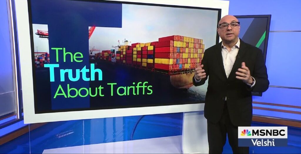 A bald man in a grey suit and dark rimmed glasses stands to the right of a large white-framed monitor which reads The Truth About Tariffs and shows a stack of shipping containers in the background.