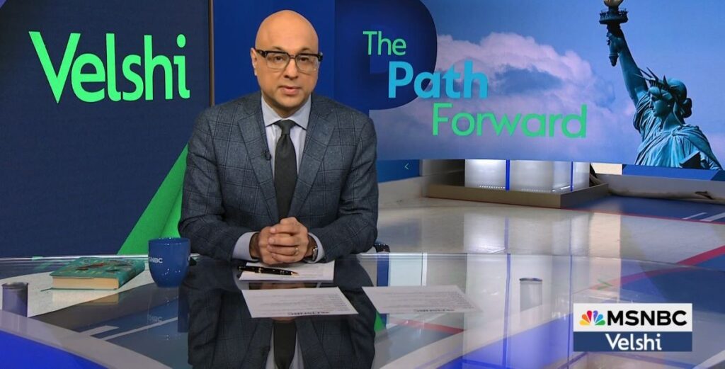 A bald man in a grey suit and dark blue tie with black rimmed glasses sites behind a large desk with a large graphic monitor behind him that reads "Velshi" to his left and to his right, "The Path Forward"