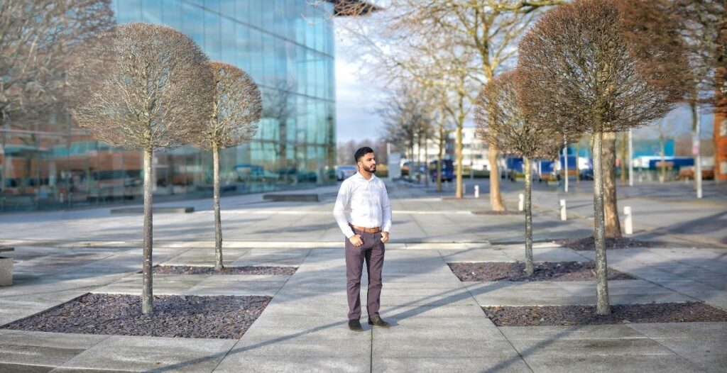 A lone man with facial hair wearing a light blue button up shirt and beige slacks stands outside an office building amongst several planted trees along a sidewalk.