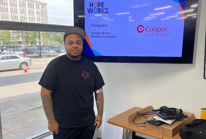 Aniage Brown, an African American 21-year-old wears a cap, has arm tattoos and wears a black t-shirt while standing in front of a screen that says "Cooper."