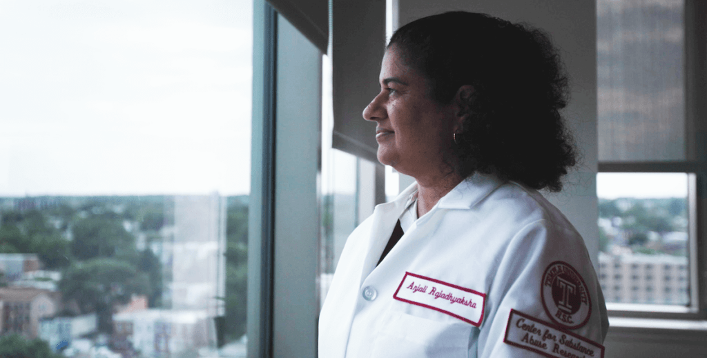 Researcher and neuroscientist Dr. Anjali Rajadhyaksha, an Indian American woman with curly shoulder length hair wearing a lab coat bearing her name looks out a window from her lab at Temple University Health.