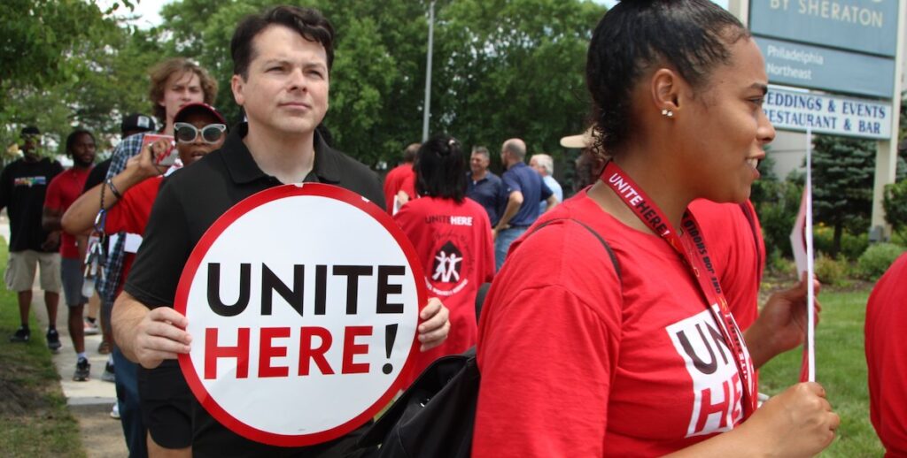 U.S. Congressman Brendan Boyle, a pale white 45-year-old man with dark short curly hair, stands while carrying a round sign bearing the words "UNITE HERE!" He is part of an outdoor procession of people — most apparently, Black women wearing red t-shirts.