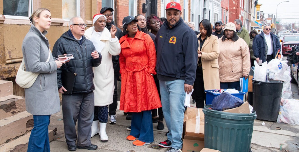 Philadelphia Mayor Cherelle Parker, an African American woman wearing a blue baseball cap, blue jeans and a red trench coat and red sneakers, stands with a group among household trash on a sidewalk in Philadelphia. Beside her is Director of Clean and Green Initiatives Carlton William, an African American man with a beard wearing a blue jacket, red cap and blue jeans.