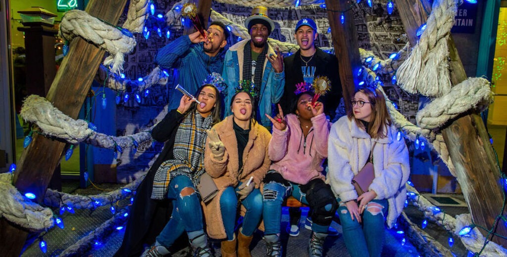 A group of seven people, Black and white, women and men, stand and sit between some kind of structure with large ropes twisted around wooden beams and blue string lights. They are wearing outdoor jackets and some are in New Year's Eve hats, looking silly.