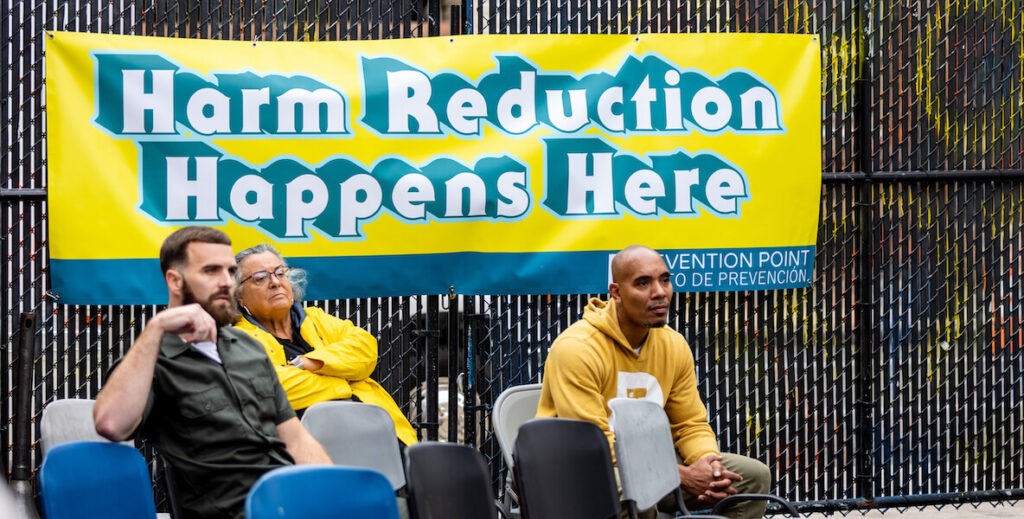 Three people sit in plastic chairs outside a fence bearing a sign that says, "Harm Reduction Happens Here." Attendees listen to guest speakers at Prevention Point’s Recover and Thrive event on Sept. 25, 2024.