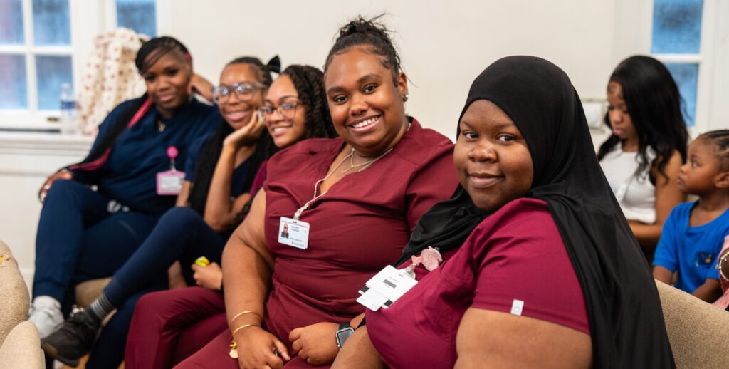 A group of young adult African American women wearing scrubs — and one a hijab — sit in chairs and smile for the camera at Hopeworks' Medical Assistant training graduation.