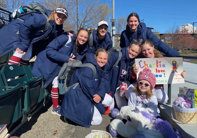 A group of young women softball players in blue team coats hold up a colorful sigh reading "Charlie we love you" while a young girl in a pink hat with a stuffed unicorn and basket of toys sits in front of them