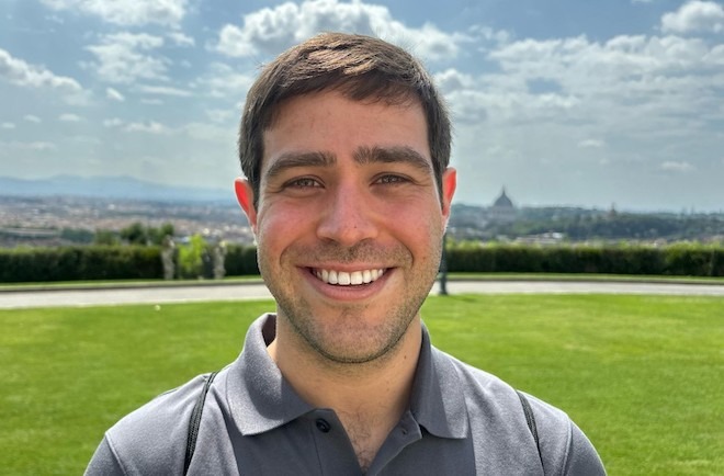 A headshot of Julian Plotnick, a white man with short brown hair, wide eyebrows and stubble wearing a polo shirt smiles while on a lawn. In the distance behind him is a city skyline. 