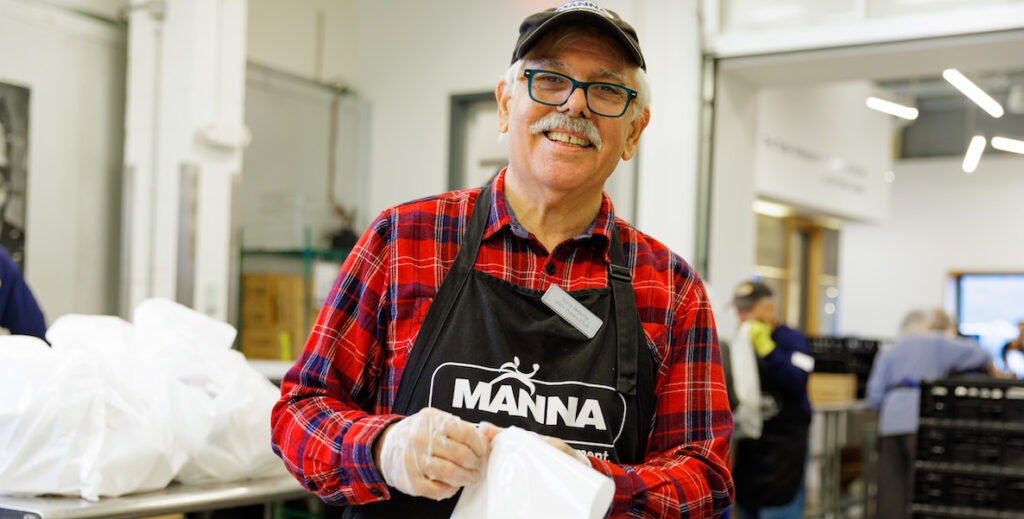 A white man with white hair, a grey and white mustache, blue-framed glasses, a cap, a red and plaid flannel shirt, black apron that says "MANNA" and safety gloves holds a bag and smiles in the MANNA warehouse.