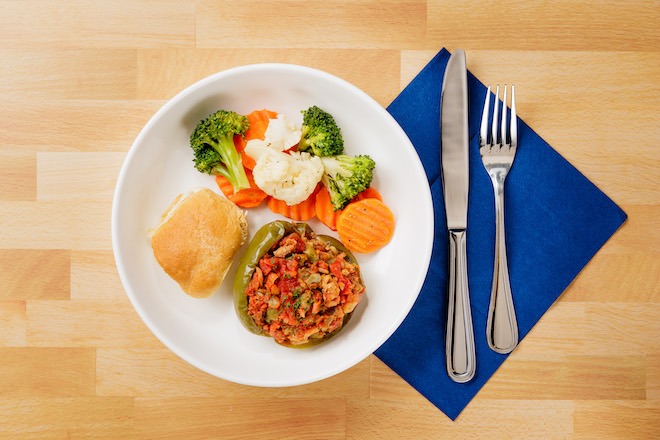 A medically-tailored meal from MANNA on a white plate includes cauliflower, broccoli and carrots, a roll, and a stuffed green pepper. Below the plate is a blue napkin. Also atop that napkin are a knife and fork, on a wooden surface.