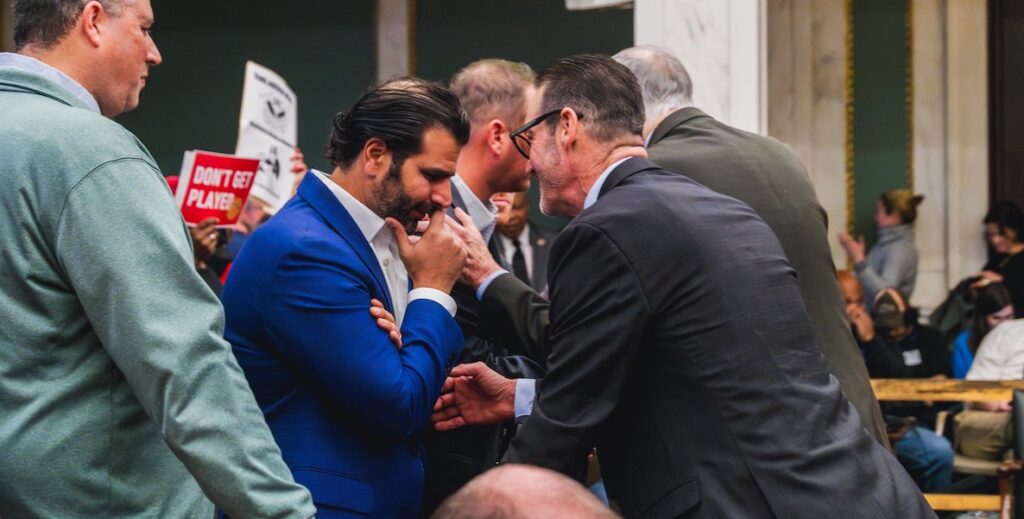 A group of people stand in City Council chambers. In the foreground stands a large white man with grey and white curly hair and a small bald spot, wearing two collared tops, while facing Mo Rushdy, an Egyptian man with slicked back black hair and a large bald spot in a bright blue suit and open-collared white shirt, looking down with his left arm crossed and right hand to his nose, listening to Councilmember At-Large Jim Harrity, a white man with short black and grey hair, a trim white beard wearing a grey suit and smiling while leaning toward Rushdy. This is a "committee of the whole" meeting of City Council about the 76 Place legislation. Behind the men, people hold signs protesting the proposed 76ers arena. One says "DON'T GET PLAYED."