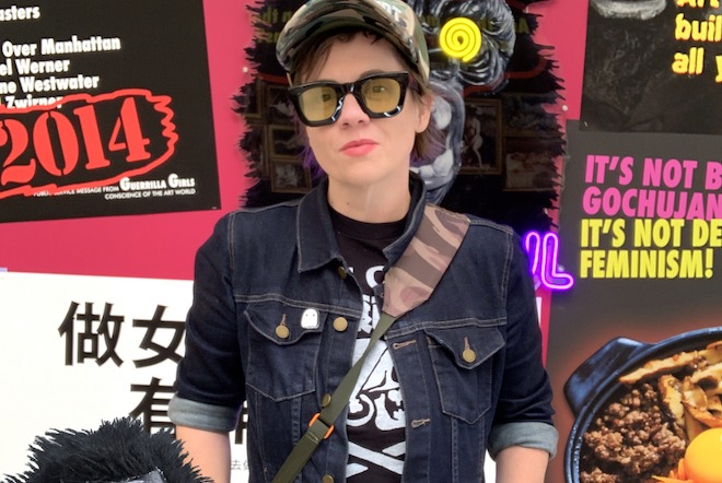 Natalie Hope MacDonald, an artist and writer and white woman with short brown hair, wearing a camo cap, large-frame glasses, a fitted jeans jacket with a ghost pin, black and white graphic shirt and camo cross-body bag stands in front of posters she created about feminism