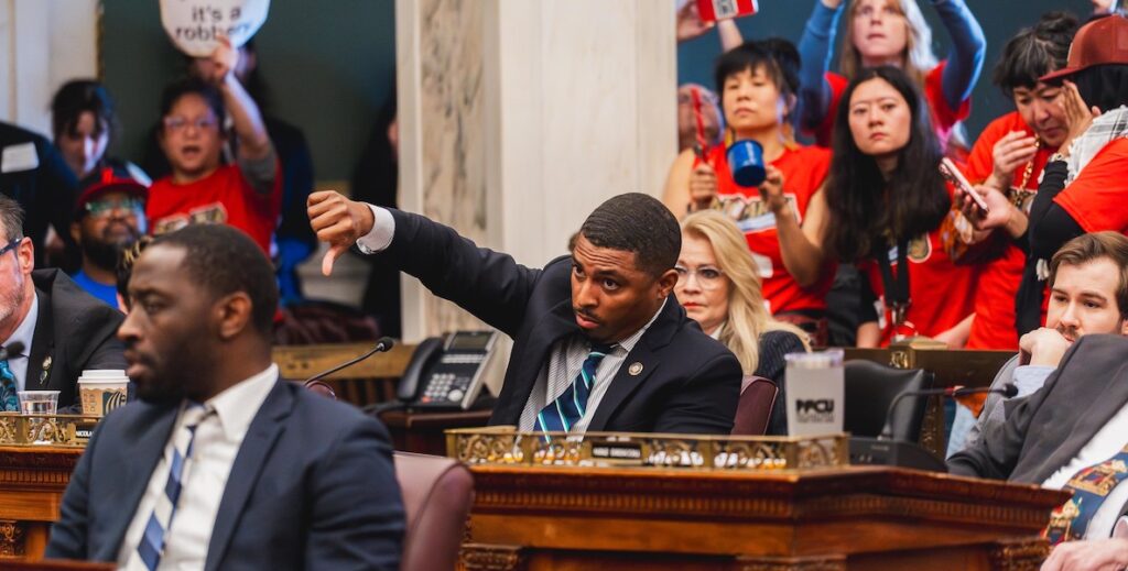 In Philadelphia City Council chambers, Councilmember Nicolas O'Rourke, a Black man with short hair wearing a dark suit, striped tie and blue collared shirt, sits behind his desk giving a "thumbs down" to proposed 76 Place legislation. In the foreground is fellow councilmember Isaiah Thomas. Behind him are anti-arena protestors.