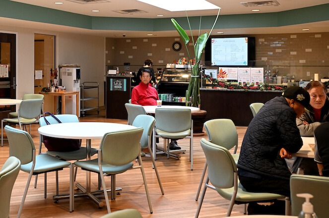 In. a large cafe setting, a lone senior, a an Asian man in a red sweater, sits at a table. Other older Asian people sit around another table. The rest of the tables appear to be empty.