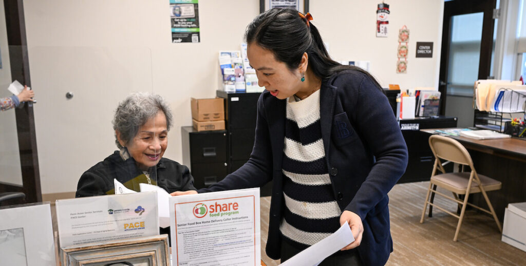 An Asian senior citizen client sits at a desk blocked off with paperwork about benefits for food and healthcare while a worker at PASSi, Penn Asian Senior Services, leans over to assist her. The worker is an Asian woman with long black hair pulled back with a clip, wearing a navy blue jacket and striped sweater. They are in an office setting.