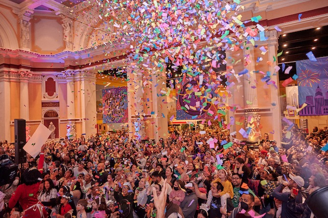 In a large, ornate room, a group of children and adults stand beneath a cloud of falling confetti on the New Year's Eve celebration at the Please Tuuch Museum.