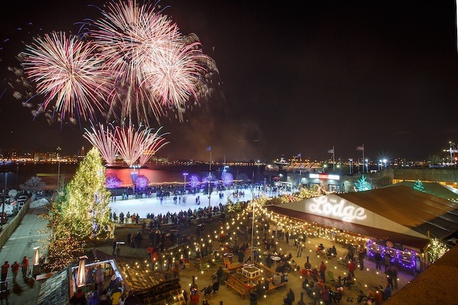 Fireworks light up the sky over the Delaware River. The aerial view displays the RiverRink outdoor ice skating rink, a large, lit-up Christmas tree, and strung white lights next to a building with a sign that says "The Lodge."