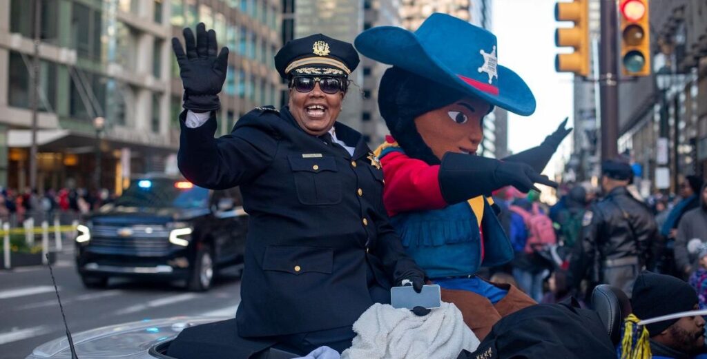 A Black woman wearing the black Philadelphia Sheriff uniform and sunglasses is sitting on a convertible next to a mascot character, waving her right hand