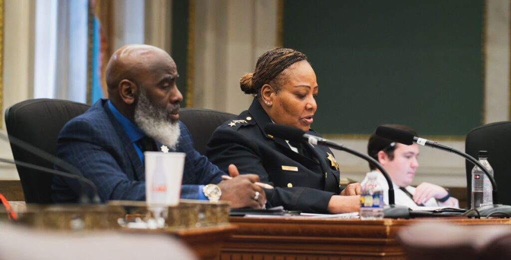 A Black woman in a black sheriff's uniform sits at a large wooden desk with a microphone before here. She is Philadelphia Sheriff Rochelle Bilal. Beside her is a bald Black man in a dark suit with a white beard