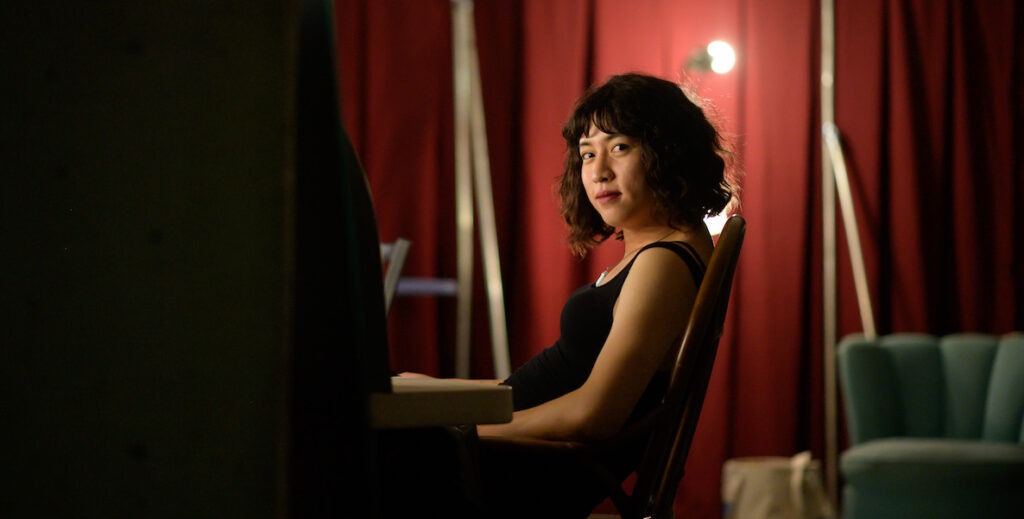 Tan Hoang, an Asian American stand-up comedian with curly black bobbed hair, wearing a black tank top, sits in a chair and looks toward the camera. Behind her is a red curtain, floor lamp and upholstered chair.