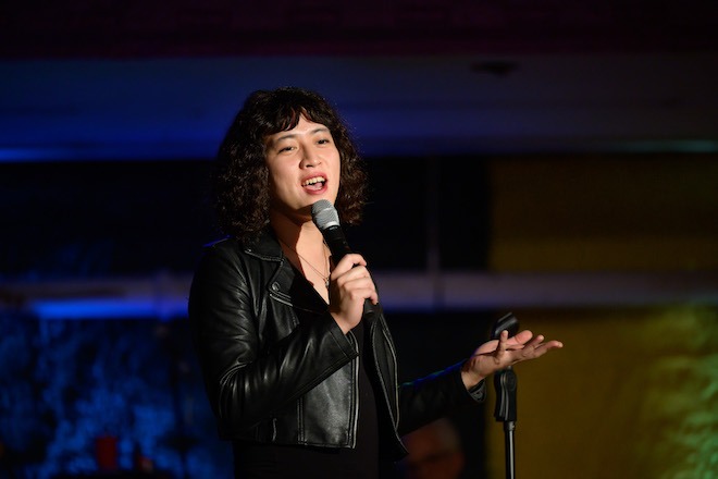 Tan Hoang, an Asian American stand-up comedian with curly black bobbed hair with bangs, wears a black leather jacket and holds a mic while talking / performing at the Bearded Ladies Cabaret.