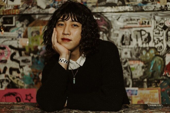 Tan Hoang, an Asian American stand-up comedian with curly black bobbed hair, wearing a black sweater over a collared white shirt, sits with her chin in her hand in front of a graffiti- art-strewn backdrop at Tattooed Mom's, a bar on South Street in Philadelphia.