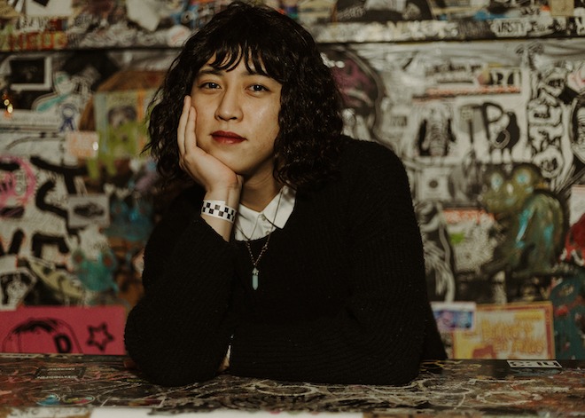 Tan Hoang, an Asian American stand-up comedian with curly black bobbed hair, wearing a black sweater over a collared white shirt, sits with her chin in her hand in front of a graffiti- art-strewn backdrop at Tattooed Mom's, a bar on South Street in Philadelphia.