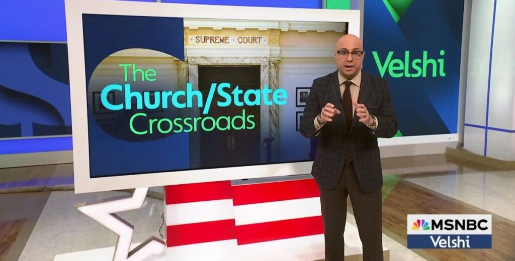 A bald man in black glasses wearing a dark plaid suit jacket and a brown tie stands to the right of a large monitor that reads "The church/state crossroads"