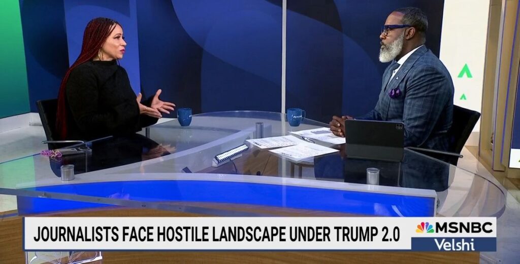 A Black woman with long hair wearing black sits across a large anchor desk on the left, while a Black man in a blue striped suit and a grey beard and blue glasses sits on the right with papers in front of him. The banner reads "Journalist face hostile landscape under Trump 2.0"