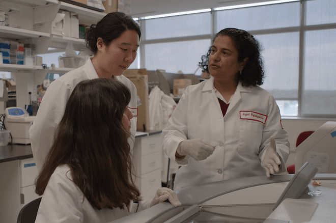 Researcher and neuroscientist Dr. Anjali Rajadhyaksha, an Indian American woman with curly shoulder length hair wearing a lab coat bearing her name appears to speak with two other women in lab coats — one seated, one standings — at her lab at Temple University Health.