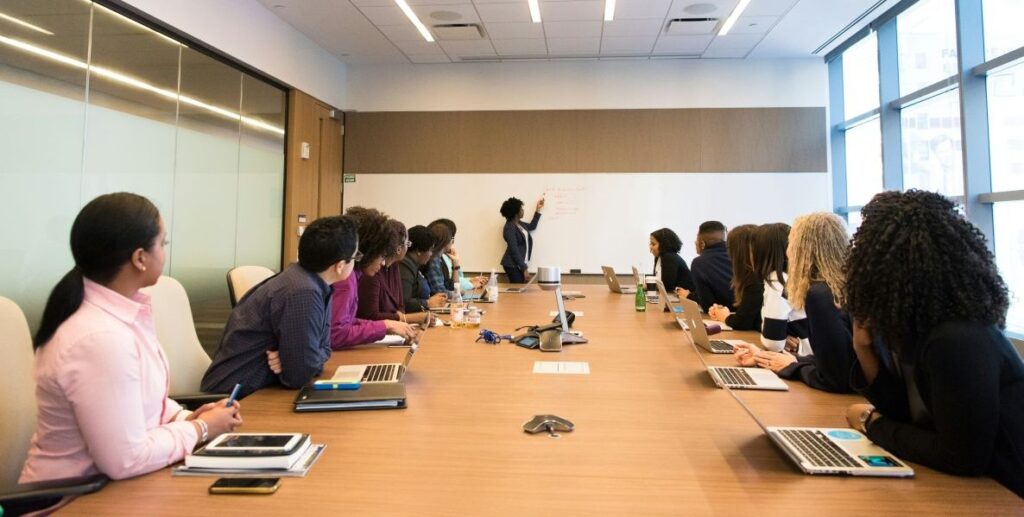 A large pine boardroom table stretches from the foreground into the background, with adults in business wear sitting along each side, each with a notepad or laptop in front of them. A woman in a dark suit stands at the white board at the head of the room, pointing with a marker