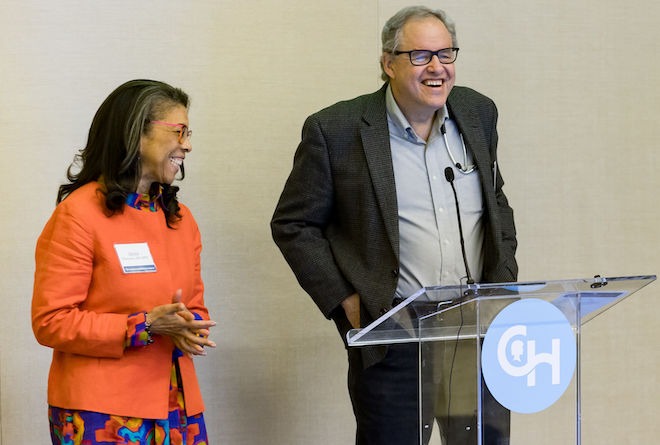 Doctors Alexis A. Thompson (left) and Stephan A. Grupp of the Children's Hospital of Philadelphia stand side by side during a presentation. Thompson, an African American woman with long, straight hair, wears glasses, a nametag, an orange jacket over a multicolored dress, stands beside Grupp, a White man with thinning hair, glasses, an unbuttoned suit jacket, button-down shirt and slacks, standing behind a clear podium with the Children's Hospital of Philadelphia logo, a large C and H. 