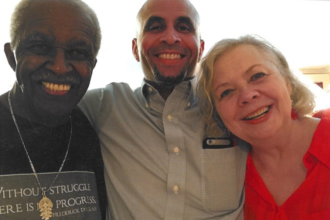 Dr. Bill McKinney, a bald African American man with a slight beard wearing a button-down shirt stands with his arms around his parents, a Black man with a white mustache wearing a black t-shirt that says "Without Struggle There Is No Progress" and a gold chain, and his mother, a white woman with blonde hair wearing a red blouse. They are all smiling.