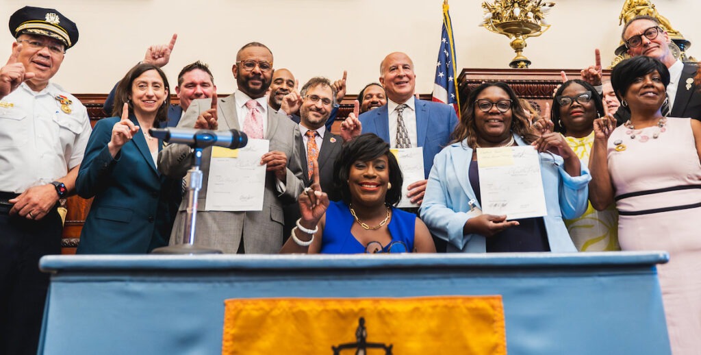 Philadelphia Mayor Cherelle Parker (center) sits at a table adorned with the Philadelphia flag. Behind her stand members of her administration, along with Police Commissioner Kevin Bethel (far left), City Councilmembers Rue Landau, Curtis Jones Jr., Mark Squilla, Kendra Brooks, Jim Harrity. The group holds up their index fingers to show "One Philly" after the mayor signs three public safety bills in September 2024.