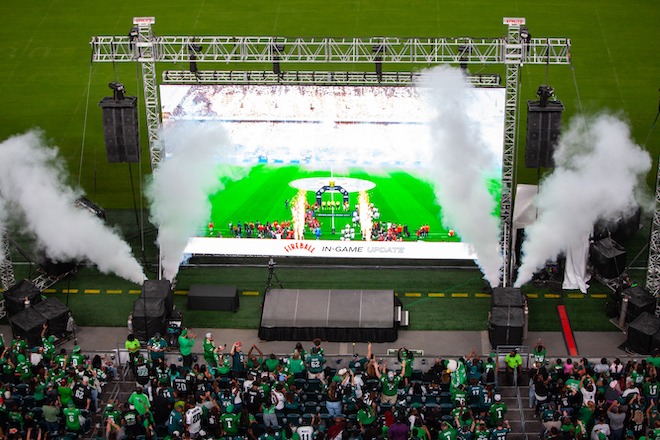 During a watch party for the Eagles-Packers game in São Paulo, Brazil, a large screen on the edge of the field at Lincoln Financial Field shows the game and features steam rising on either side. Facing the screen are stadium rows of Eagles fans who've paid to come to watch the game.