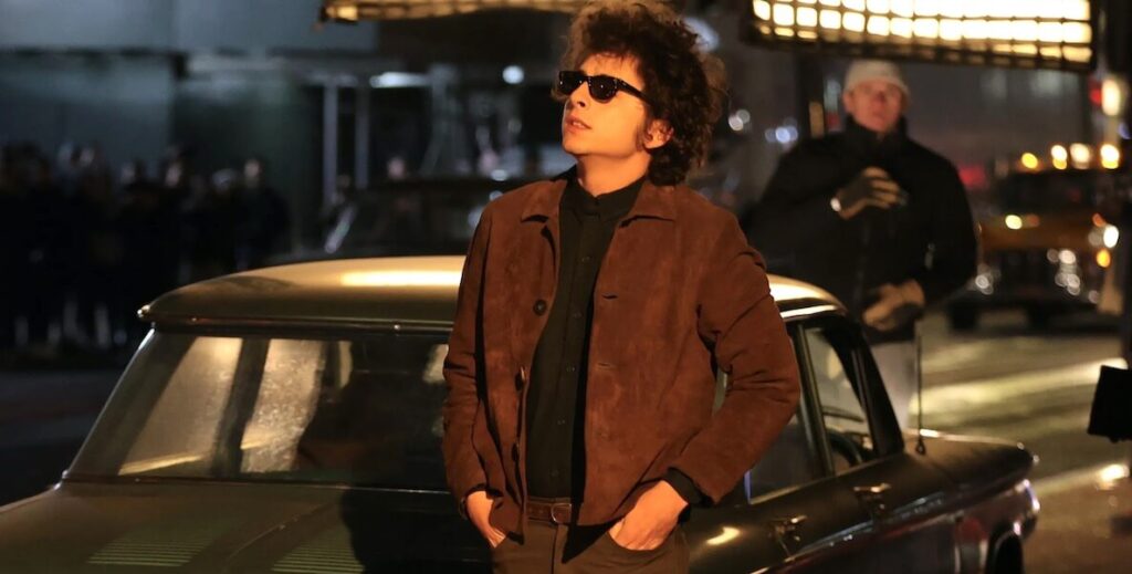 Timothée Chalamet, a White man with dark brown shaggy curly hair, wearing Wayfarer-style dark sunglasses, a collared brown jacket and black shirt, stands with his hands in his pants' pockets, looking up at a marquee. He portrays musician Bob Dylan, standing outside a theater, leaning against a car in a street, in A COMPLETE UNKNOWN.