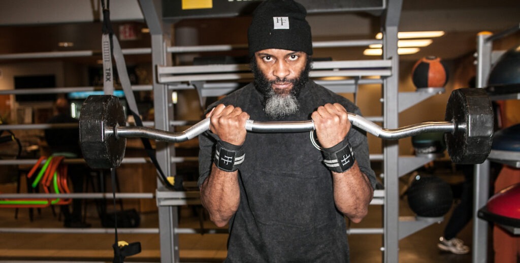 Jamar Blackshear, a Black man with a black and white beard, lifts a weight and looks into the camera. He is wearing a Halal Fitness black beanie, a black t-shirt and wristbands.