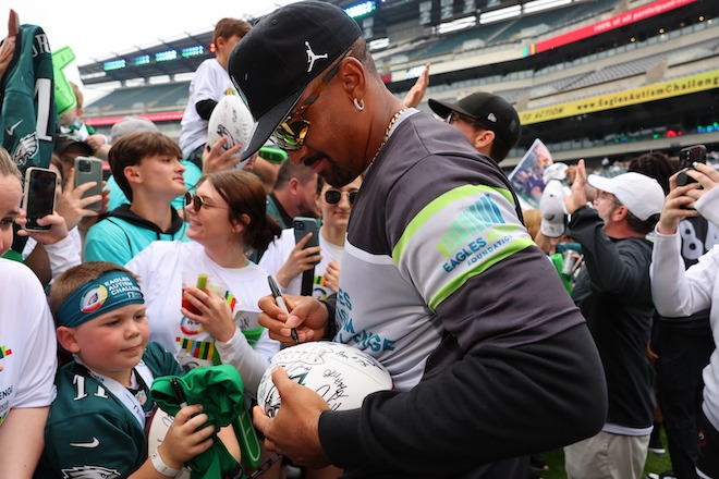 Jalen Hurts, the Philadelphia Eagles quarterback, wearings a black Nike cap, diamond hoop earring in his left earlobe, sunglasses, a wide chain and an technical Eagles Autism Foundation shirt, signs a white Eagles football for a child wearing an Eagles jersey and Eagles Autism Foundation headband. They are in a crowd of people on the field at Lincoln Financial Field football stadium.