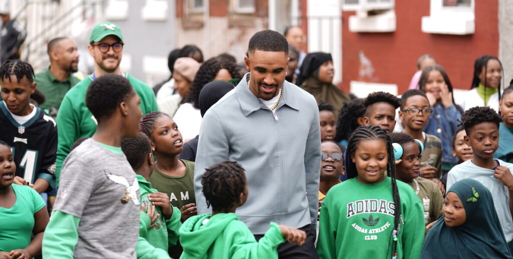 Philadelphia quarterback Jalen Hurts, a Black 25-year-old with a sweet fade and a zip-up grey jacket, walks in Philadelphia with mostly Black students from Edward Gideon School, all wearing Eagles green.