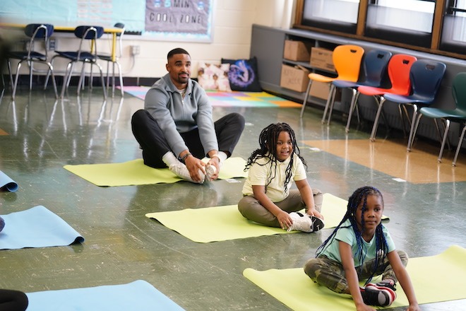 Eagles Quarterback Jalen Hurts, a Black man wearing a grey zip-up sweater, black pants and white socks and sneakers, sits on a yoga mat behind two elementary school students, both African American girls with long braids wearing short-sleeve shirts pants and sneakers, also sitting on yoga mats in badhakonasana in a classroom setting.