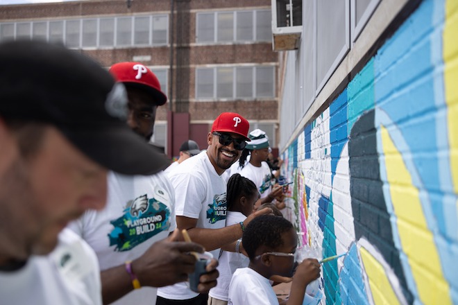 Philadelphia Eagles Quarterback Jalen Hurts, an African American man wearing a red Phillies cap, white Girard Elementary t-shirt, and dark sunglasses, stands among students to paint an outdoor mural at Stephen Girard Elementary School in Philadelphia.