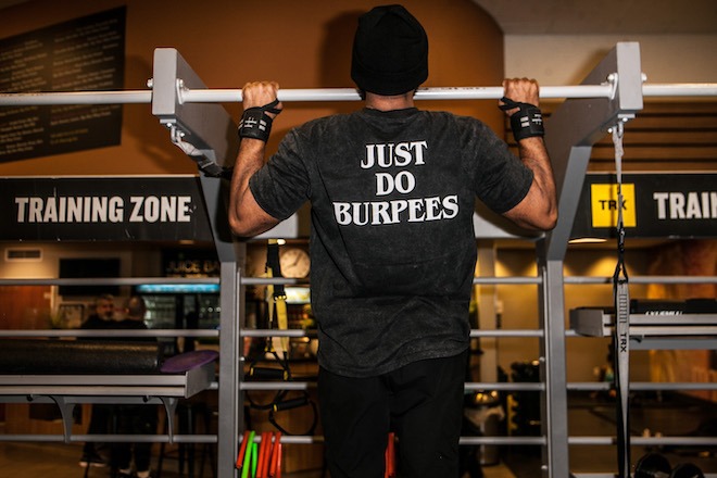 Jamar Blackshear, a Black man, does a pull-up in a gym with banners that say "TRAINING ZONE." Blackshear wears a black beanie, wristbands, pants and a t-shirt that reads "JUST DO BURPEES." His back is to the camera.