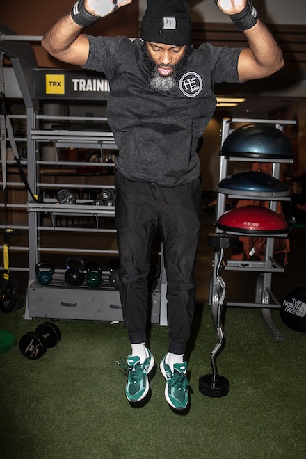 Jamar Blackshear, a Black man with a black and white beard, jumps while doing a burpee in a gym. He is wearing a Halal Fitness black beanie, a black t-shirt and wristbands and green New Balance sneakers.