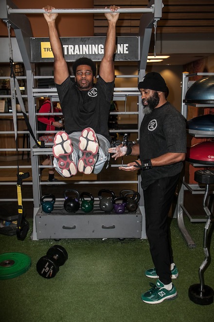 Jamar Blackshear, a Black man with a black and white beard, wearing all black and green New Balances, trains a client who is hanging from a poll and lifting his legs to 90 degrees.