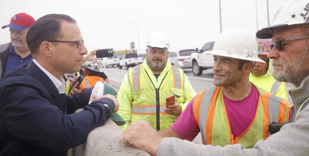 PA Governor Josh Shapiro, a white man with dark brown short hair and glasses wearing a navy jacket, speaks to workers in safety vests and hard hats on the edge of a just-repaired I-95 in June 2023.